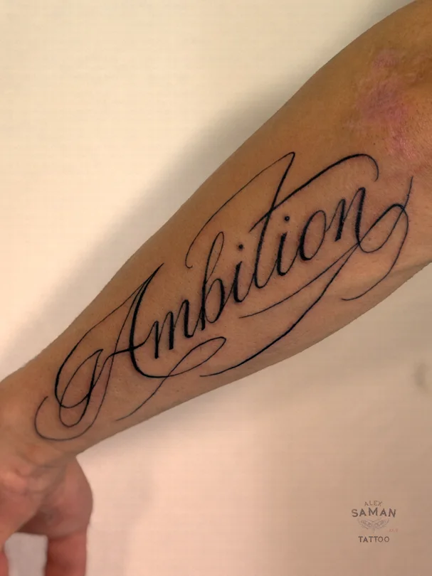 Tatouage du mot Ambition en écriture calligraphique, avec de longues courbes sur l’avant-bras.