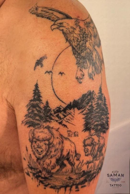 Tatouage sur le haut du bras associant un aigle en vol, un bison, des montagnes et des sapins.