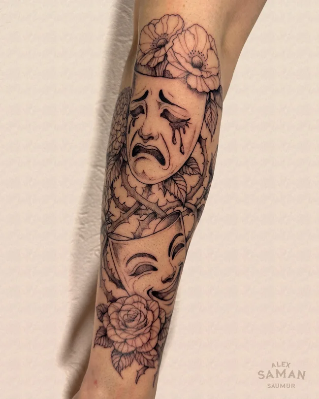 Tatouage de deux masques de théâtre, l’un triste et l’autre souriant, entourés de fleurs.
